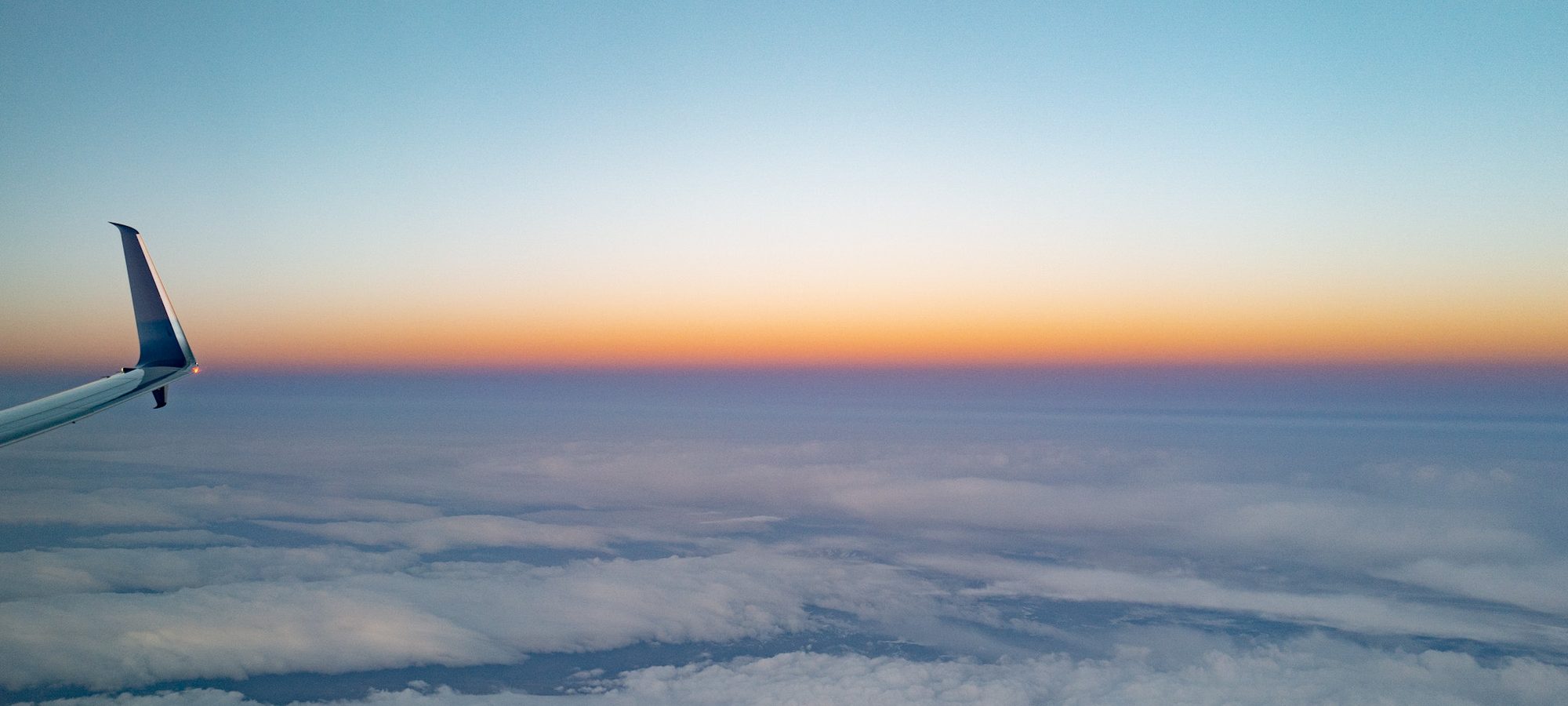 A winglet above a sea of cloud, the horizon going from orange to blue, photographed from the window seat.