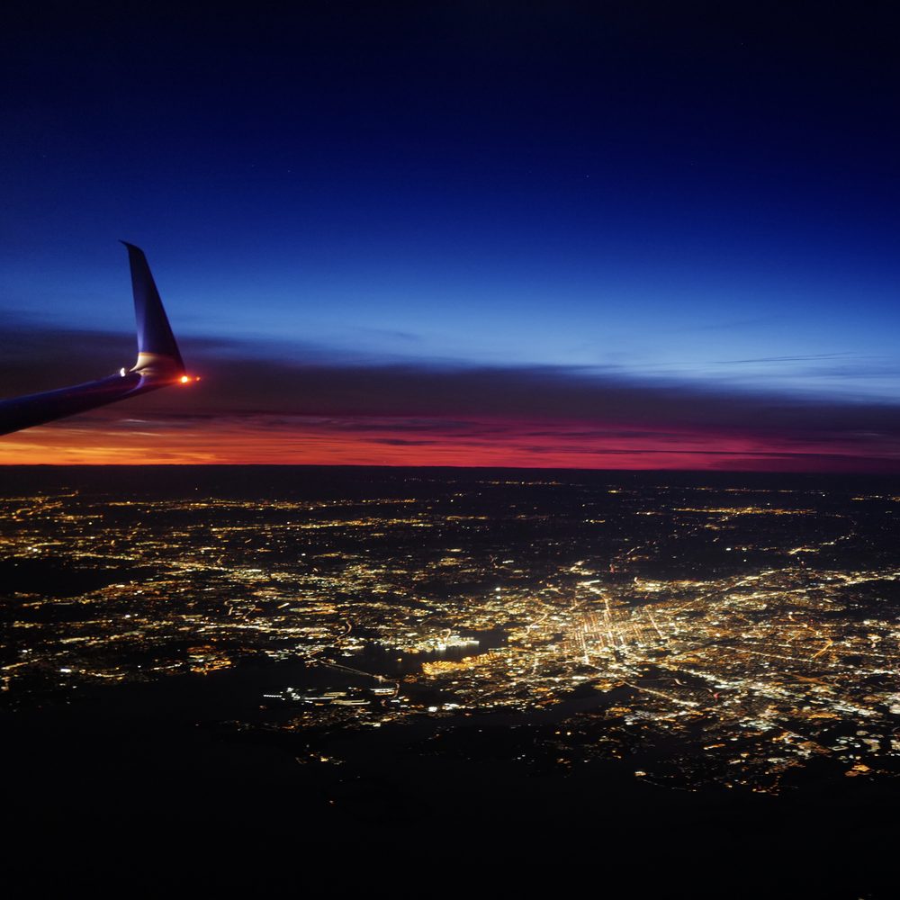 City lights below a winglet at dusk, under a red band on the horizon.