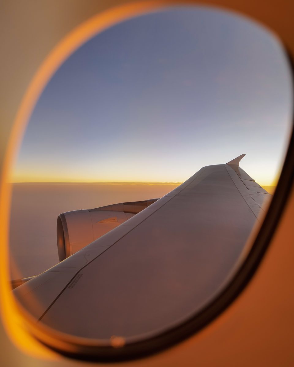 Golden light on an oval aircraft window framing a wing and the clouds below.